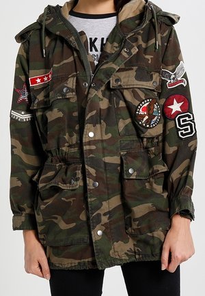 Osoba ubrana w kurtkę moro z wieloma naszywkami, w tym gwiazdami, orłem oraz literą "S" na rękawach i kieszeniach na piersi.