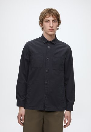 Jeune homme aux cheveux bouclés porte une veste noire boutonnée avec deux poches poitrine et un pantalon marron sur un fond blanc uni.