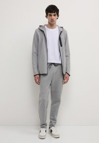 Junger Mann steht gegen eine weiße Wand und trägt graue Jogginghosen, eine graue Kapuzenjacke mit Reißverschluss, ein weißes T-Shirt und weiße Sneakers mit schwarzen Details.