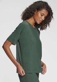 Sieva ar cirtainiem matiem, valkājot tumši zaļu ribotu īsroku kreklu un saskaņotas bikses, turot krekla apakšmalu.