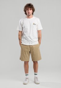 Valkoinen puuvillapaita, jossa on "Escape to Nature" -grafiikka, yhdistettynä khaki-shortsien kanssa. Asusteita täydentävät valkoiset lenkkarit ja vihreäväriset raidalliset sukat.