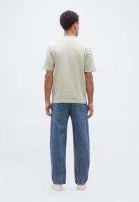 Άνδρας που στέκεται με την πλάτη γυρισμένη, φοράει ανοιχτό μπεζ πουκάμισο με κοντά μανίκια, μπλε τζιν με πίσω τσέπες και λευκά αθλητικά παπούτσια σε απλό φόντο.