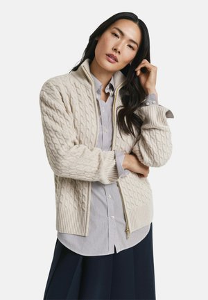 Vrouw met lang donker haar, gekleed in een beige kabelgebreide ritstrui over een gestreepte blouse en een donkere rok, poserende met haar hand nabij haar gezicht.