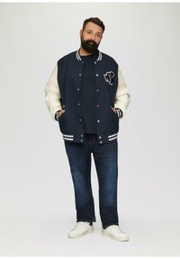 Veste de varsity bleu marine avec manches crème, rayures blanches et logo NY. Associée à un jean foncé et des baskets blanches.