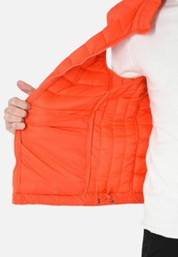 Gilet arancione imbottito con motivo trapuntato, chiusura con zip, taschini interni e cordoncino all'orlo. Tessuto leggero e liscio.