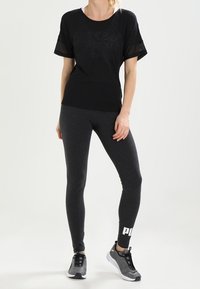 Svart atletisk t-shirt med meshärmar, tillsammans med mörkgrå leggings med en vit logotyp, och grå atletiska skor med svarta accenter.