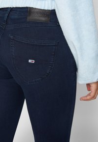 Vaqueros de denim azul oscuro con una superficie texturizada, que presentan un bolsillo trasero acentuado por un pequeño logo bordado en rojo, blanco y azul.