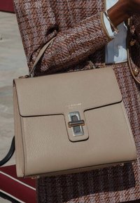 Beige Lederhandtasche mit glatter Textur, ausgestattet mit einem silbernen Metallverschluss und einem strukturierten Design. Kombiniert mit einer gemusterten Jacke.