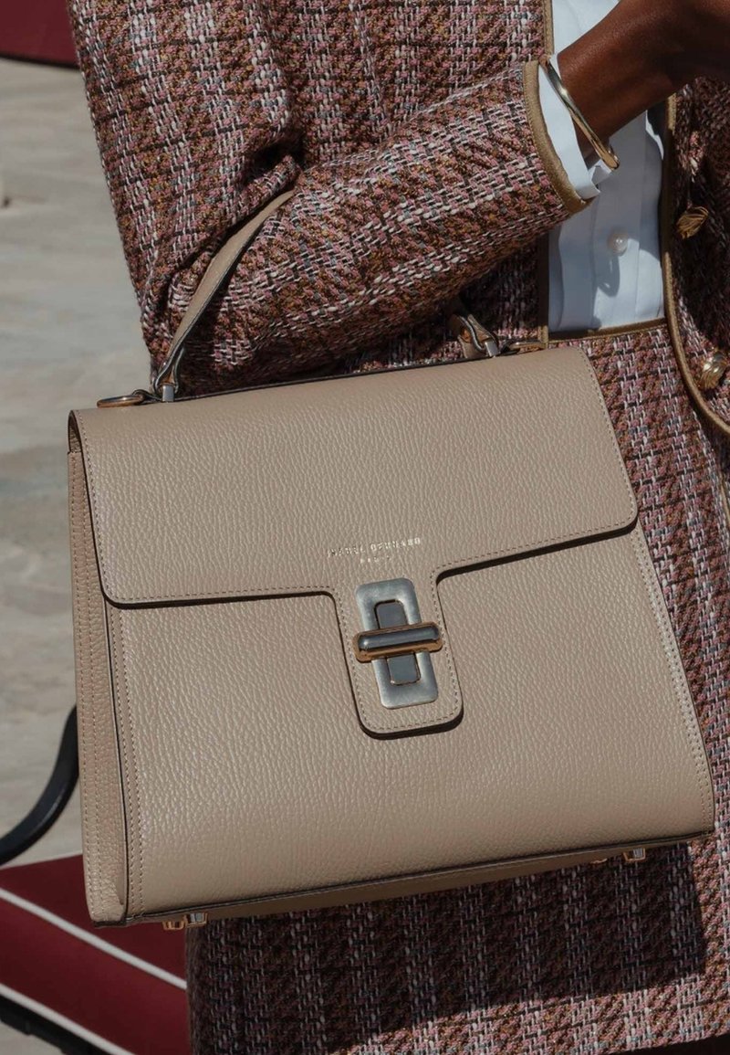 Beige Lederhandtasche mit glatter Textur, ausgestattet mit einem silbernen Metallverschluss und einem strukturierten Design. Kombiniert mit einer gemusterten Jacke.