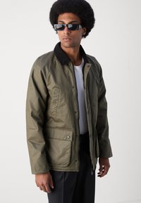 Chaqueta bomber verde oliva con cuello de pana negra, cierre de cremallera y bolsillos delanteros con parche. Textura suave y corte relajado.