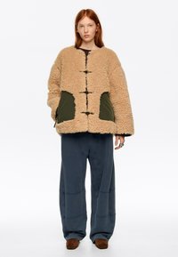 Mujer de cabello rojo que lleva una chaqueta beige de piel sintética sobredimensionada con bolsillos verdes, pantalones oscuros y sueltos, y zapatos marrones, de pie sobre un fondo blanco.