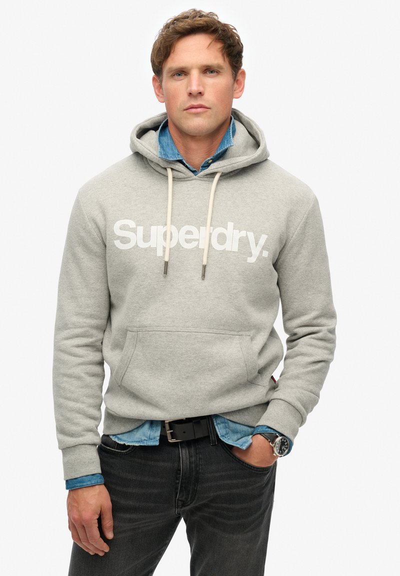 Un sweat à capuche gris avec un logo "Superdry" blanc, des cordons de serrage et une poche avant. Porté par-dessus une chemise en denim bleu et associé à un jean foncé.