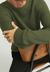 Zelený žebrovaný pletený svetr s kulatým výstřihem, který má texturovanou tkaninu a středně dlouhé rukávy, nošený při sezení na dřevěné židli.