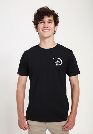 Jeune homme aux cheveux bouclés souriant, portant un t-shirt noir avec un logo "D" stylisé en blanc sur le côté gauche de la poitrine, pantalon beige, debout.