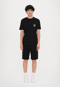 Svart bomullst-skjorte og matchende shorts. T-skjorten har en liten logo i hvitt; modellen har på seg hvite sneakers med ankelsokker. Minimalistisk stil.