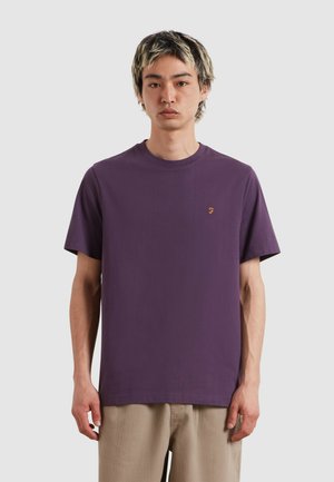 Giovane uomo con capelli corti a due tonalità, che indossa una semplice maglietta viola e pantaloni beige, in piedi davanti a uno sfondo grigio chiaro.