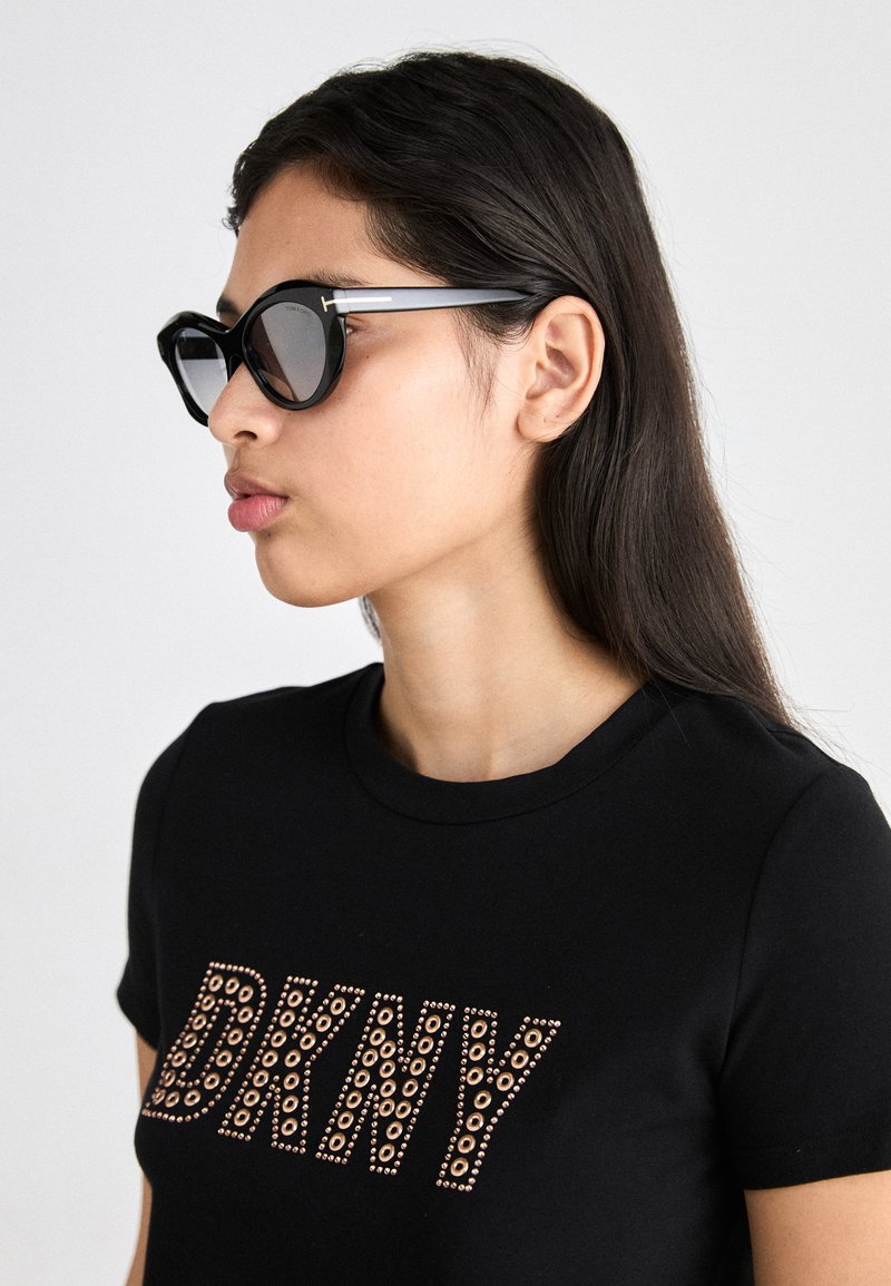 Sorte solbriller med dristige geometriske rammer, accentuert med sølv detaljer på bøylene, båret med en svart t-skjorte som har en naglet DKNY-logo.