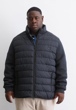Veste matelassée en marine foncé avec texture matelassée, col montant, zips frontaux et manches en tissu solide. Stylée sur une chemise en denim.