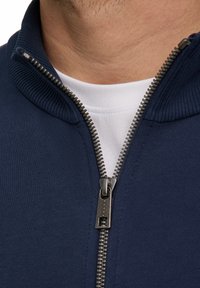 Giacca zip-up blu navy con colletto a costine, cerniera argentata e una maglietta bianca visibile all'collo. Dettaglio in tessuto testurizzato.