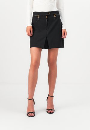 Black mini skirt with a fitted waist, gold zipper accents, a front slit, and a smooth texture. Worn with black strappy heels.