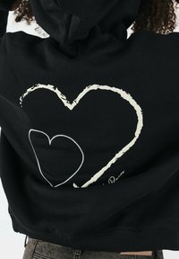 Felpa nera con cappuccio caratterizzata da una grande grafica a forma di cuore bianco sul retro, con una morbida texture del tessuto e polsini e orlo a coste per un look aderente.