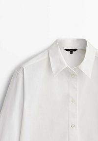 Chemise blanche boutonnée avec un col classique, des coutures visibles sur l'épaule et trois boutons au centre devant.
