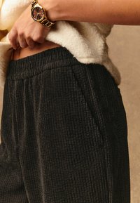 Zwarte corduroy broek met een gestructureerd, ribbelachtig ontwerp. Voorzien van een elastische tailleband en zijzakken. Gekoerd met een crème trui en een gouden horloge.