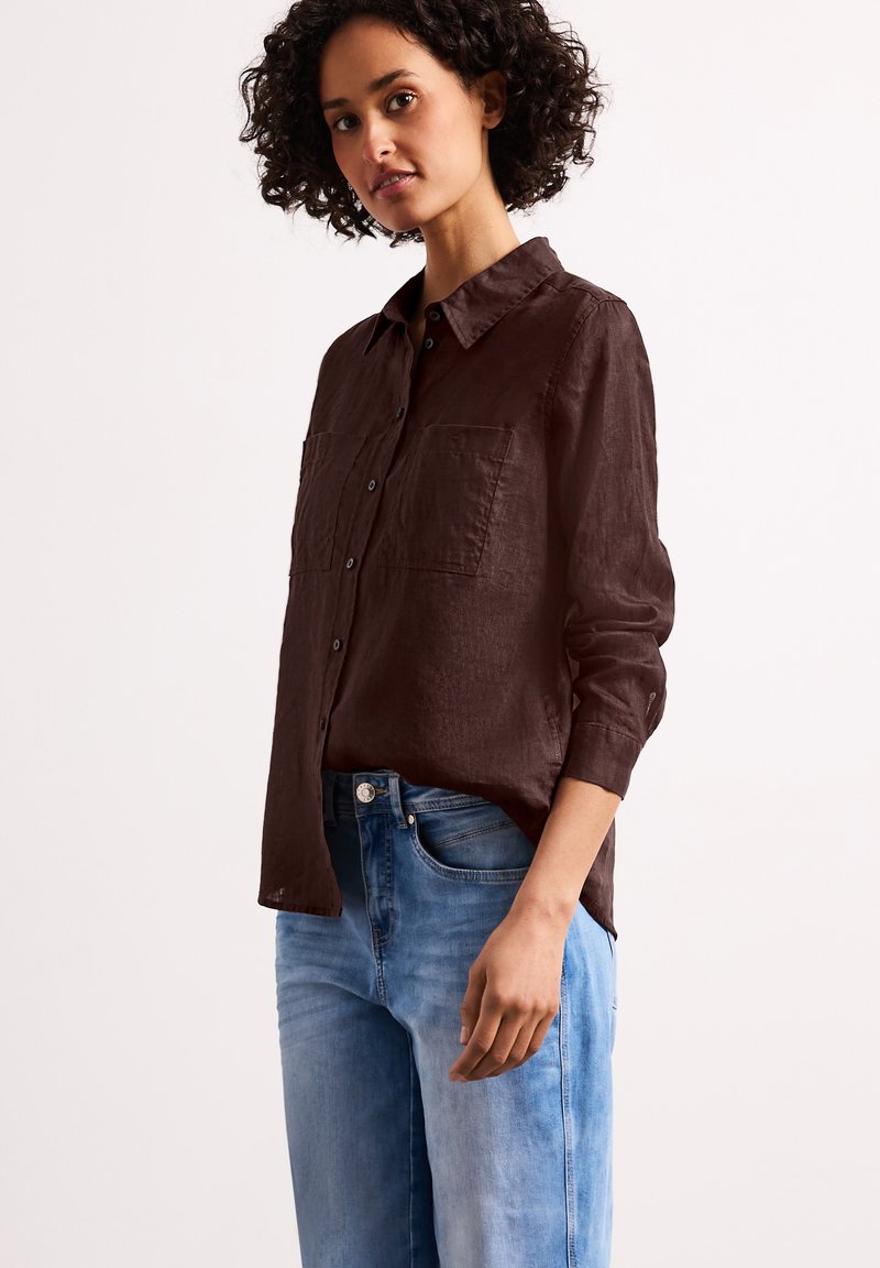 Femme aux cheveux bouclés portant une chemise marron foncé boutonnée et un jean bleu clair, debout devant un fond clair uni.