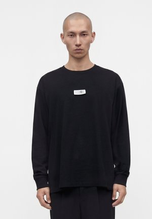 Homme à la tête rasée portant un t-shirt noir à manches longues avec une petite étiquette blanche rectangulaire au centre de la poitrine.