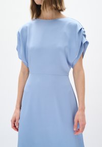Un vestito azzurro chiaro con un tessuto testurizzato, ampia scollatura a barca e maniche corte drappeggiate, caratterizzato da una vita aderente e una gonna fluente.