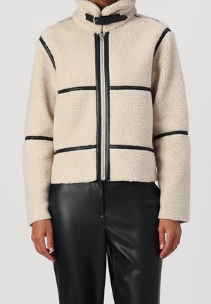 Beige sherpa-jas met zwarte biezen, ritssluiting aan de voorkant en gesp aan de kraag, gedragen met zwarte leren broek door een staand persoon.