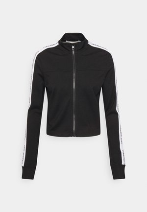 Chaqueta negra con cremallera y cuello alto, con rayas blancas a lo largo de las mangas que presentan el texto repetido "Calvin Klein Jeans".