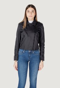 Giacca nera corta con finitura lucida, cerniere asimmetriche e colletto classico; abbinata a jeans blu aderenti.