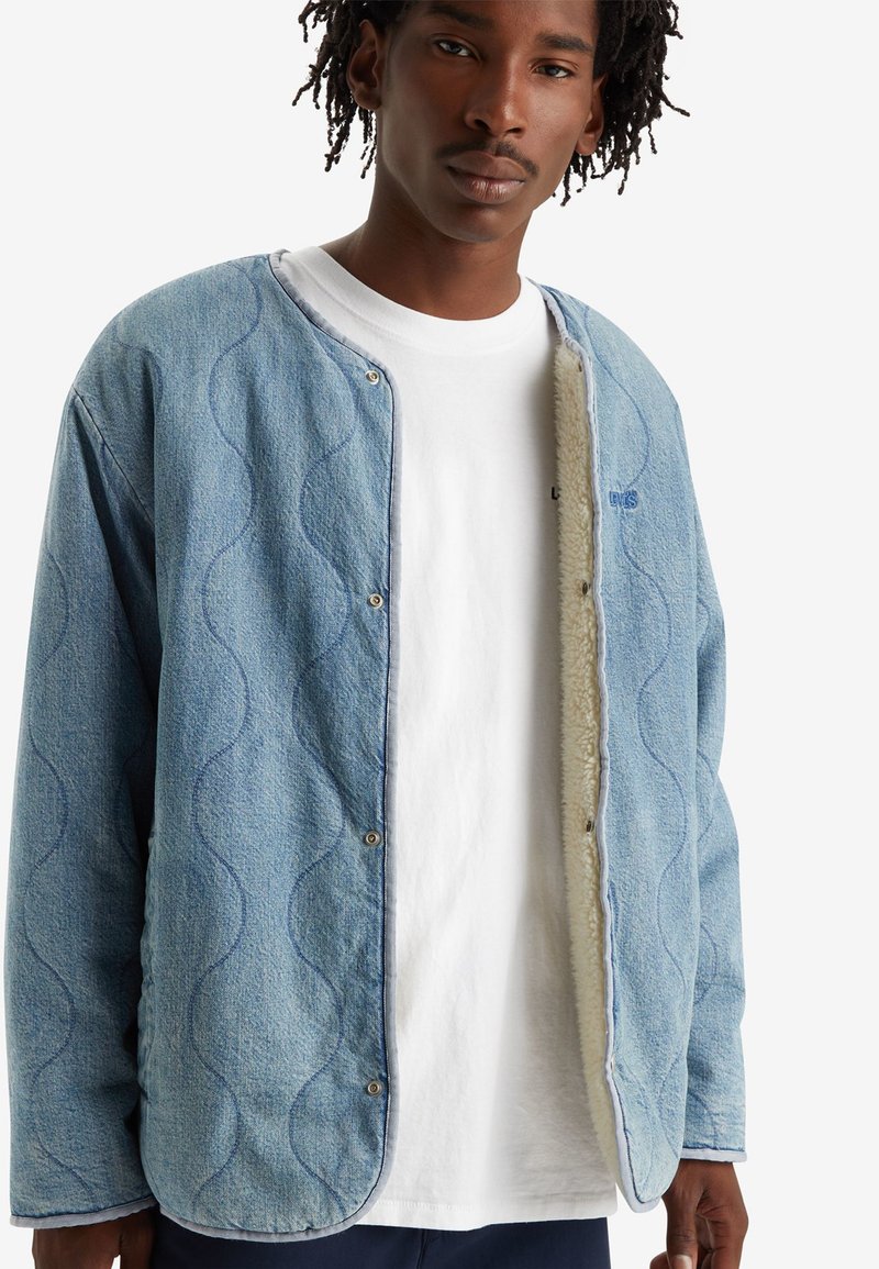 Chaqueta de denim azul claro con textura ondulada, con botones en la parte delantera y un forro suave de color crema. Presenta un cuello redondo y una mínima marca.