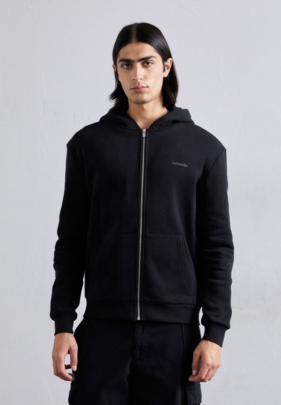 Black zip-up hoodie made of soft fabric, featuring a front pocket, ribbed cuffs, and minimal branding on the chest.