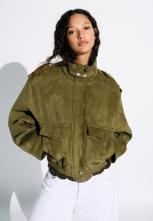 Žena s dlhými tmavými vlasmi má na sebe nadrozmernú olivovo zelenú semišovú bundu s vysokým golierom a biele nohavice na bielom pozadí.