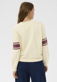 Sudadera de color crema con mangas largas, que presenta rayas horizontales en burdeos en las mangas y una cintura acanalada. Textura de tela suave.
