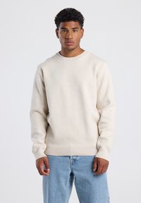 Beige knit sweater with crew neckline and long sleeves, featuring ribbed cuffs and hem. Worn over light blue denim jeans.