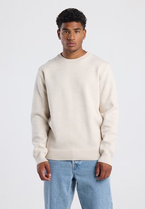 Beige gebreide trui met ronde hals en lange mouwen, voorzien van geribbelde manchetten en zoom. Draagover een lichtblauwe spijkerbroek.
