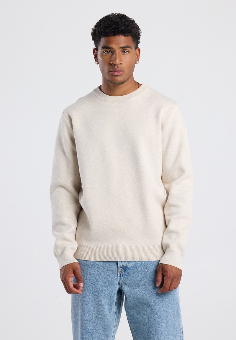 Beige knit sweater with crew neckline and long sleeves, featuring ribbed cuffs and hem. Worn over light blue denim jeans.