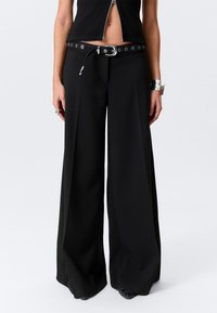 Pantalon noir à jambes larges en tissu lisse et texturé. Doté d'une ceinture à boucle métallique avec œillets. Coupe ajustée, longueur totale.