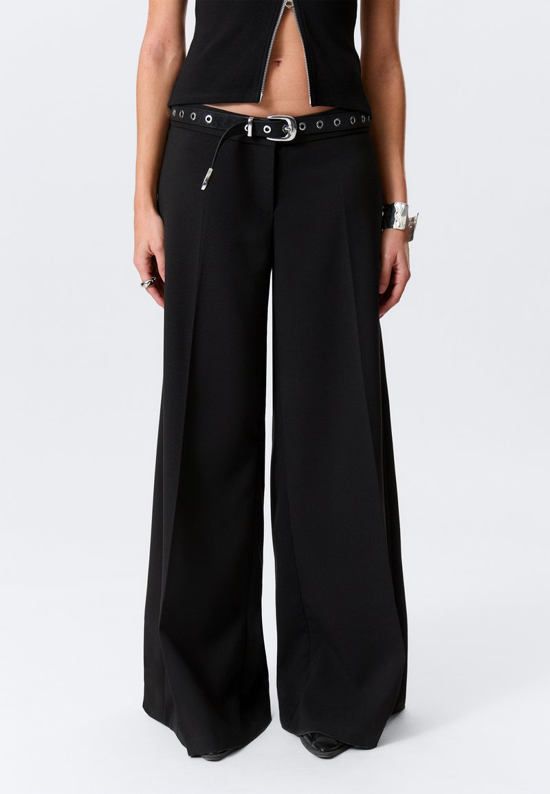 Pantalon noir à jambes larges en tissu lisse et texturé. Doté d'une ceinture à boucle métallique avec œillets. Coupe ajustée, longueur totale.