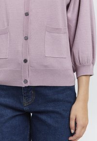 Lavendelfärgad cardigan med en texturerad stickning. Den har nedcuttade ärmar, två framfickor och sex svarta knappar längs framsidan.