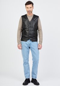 Schwarze Lederweste mit sechs Knöpfen und V-Ausschnitt, getragen über einem beigefarbenen Langarm-Shirt, kombiniert mit hellblauen Jeans und schwarzen Stiefeln.