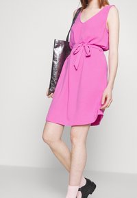 Robe rose sans manches avec un col en V et un détail de ceinture nouée, en tissu texturé. Associée à un sac fourre-tout métallique et des chaussures à plateforme noires.