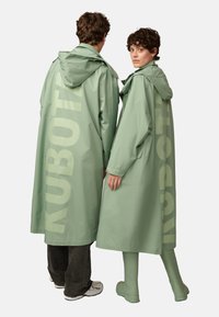 Green waterproof coats with a hood, featuring large "KUBOT" text on the back, worn over casual pants and green boots.