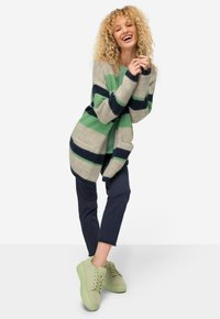 Randig cardigan i grön, marinblå och beige, tillverkad av mjukt material, med öppen framsida och två framfickor. Matchad med marinblå byxor och ljusgröna sneakers.
