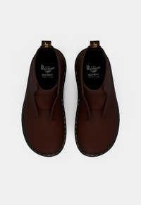 Zapatos marrones tipo slip-on con una textura de cuero suave, costuras amarillas y una suela de goma negra. Incluyen plantillas con logotipo y tiradores.