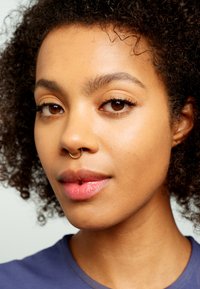 Zbliżenie na twarz z kręconymi czarnymi włosami, miodowo-brązowymi oczami, złotą pierścionkiem w septum i gładką, jasnobrązową skórą. Ubrana w niebieską koszulę.