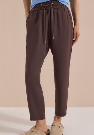 Personne portant un pantalon marron à taille élastique avec cordon, les mains dans les poches et des sandales beige à enfiler sur un fond neutre.